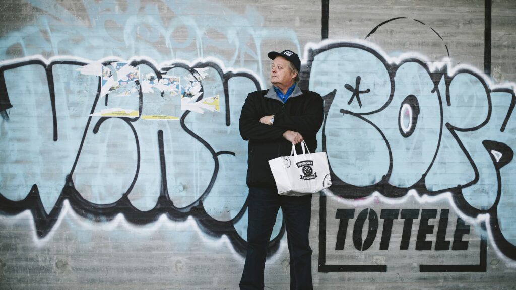 Seppo Räty poseeraamassa graffiteilla maalatun betoniseinän edessä Helsingissä Kaura Jäärä kampanjassa. Mainostoimisto Halo | Helsinki x Viipurilainen Kotileipomo